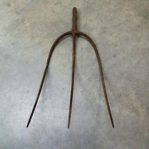May include: A rusty, antique pitchfork with three curved tines and a handle. The metal tool is designed for agricultural use, likely for turning hay or other farm tasks. The pitchfork is set against a neutral gray background.