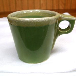 May include: A green ceramic mug with a unique handle design. The mug has a rounded body and a handle with a circular opening. The rim of the mug is a lighter shade of green. The mug is sitting on a white surface.