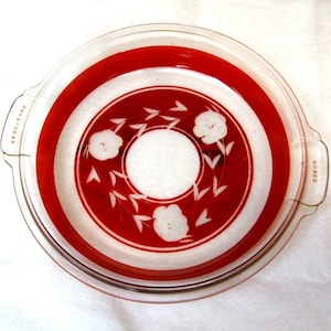 May include: A clear glass casserole dish with a red and white floral design. The dish has a circular shape with a red band around the edge and white floral accents. It has two handles and is likely vintage.