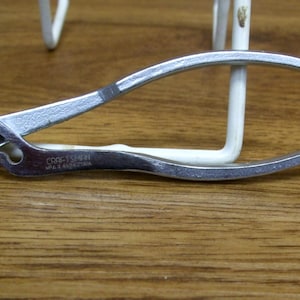 May include: A silver Craftsman slip-joint pliers with a curved handle and serrated jaws. The pliers are lying on a wooden surface, with a white metal object in the background.