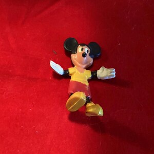 May include: A small plastic figurine of Mickey Mouse wearing a yellow shirt and red shorts. He is standing with his arms outstretched and his feet slightly apart.