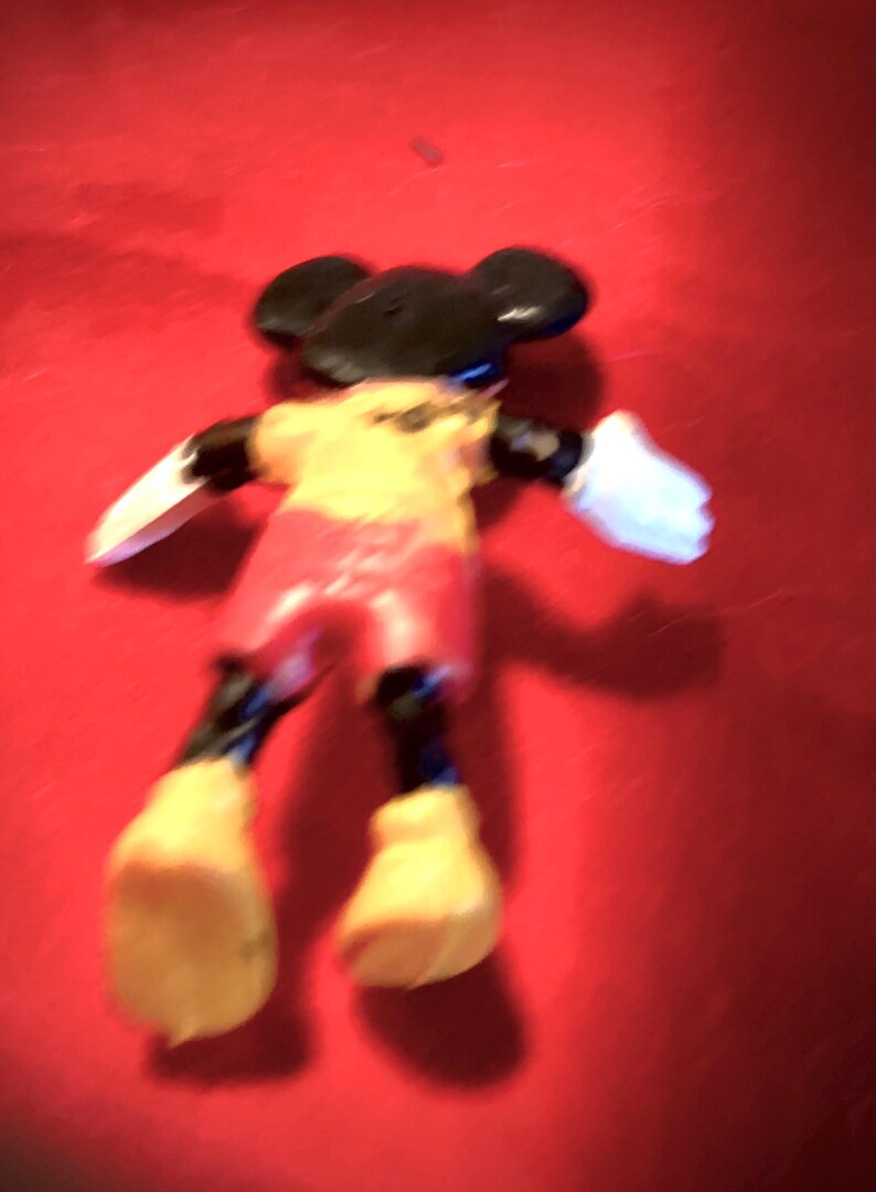May include: A black and yellow plastic Mickey Mouse figurine with red shorts and white gloves. The figurine is lying on its back on a red surface.