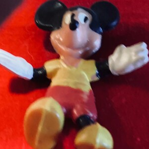 May include: A yellow, red, and black plastic figurine of Mickey Mouse. Mickey is wearing a yellow shirt and red shorts. He is smiling and has his arms outstretched.