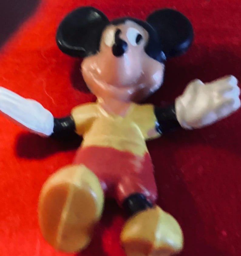 May include: A yellow, red, and black plastic figurine of Mickey Mouse. Mickey is wearing a yellow shirt and red shorts. He is smiling and has his arms outstretched.