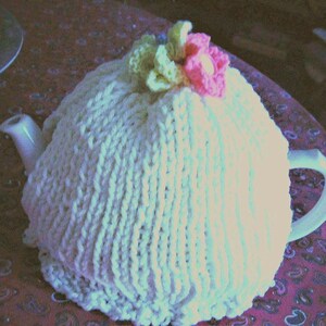May include: A white ceramic teapot covered with a cream-colored knitted tea cozy. The cozy is adorned with a light green and pink flower on top. The teapot is on a patterned tablecloth.