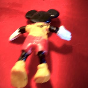 May include: A black and yellow plastic Mickey Mouse figurine with red shorts and white gloves. The figurine is lying on its back on a red surface.