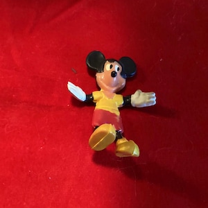May include: A small plastic figurine of Mickey Mouse wearing a yellow shirt and red shorts. He is standing with his arms outstretched and his feet slightly apart.