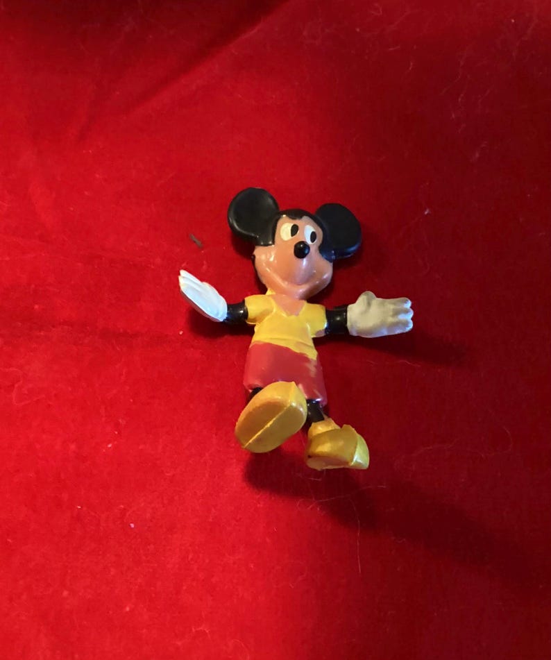 May include: A small plastic figurine of Mickey Mouse wearing a yellow shirt and red shorts. He is standing with his arms outstretched and his feet slightly apart.