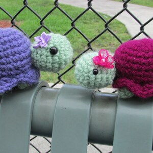 May include: Two crocheted turtle toys, one purple and one green, with pink and purple flowers on their heads. The turtles are sitting on a gray metal fence.