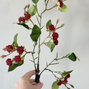 May include: A faux berry stem with dark red berries, green leaves, and small pink buds. The stem is held by a hand against a white background. The text "Dawn Renee's Designs" is visible on the stem.