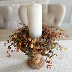 May include: A white pillar candle sits in a wooden pedestal holder, surrounded by a ring of artificial foliage with small leaves in shades of green, yellow, and red. The text Dawn Renee's Designs is visible on the foliage. The base is round.