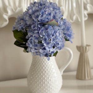 May include: A white ceramic pitcher with a textured, dotted surface holds a bouquet of blue hydrangeas. The flowers have green leaves and are arranged in a decorative vase. The text "Dawn Renee's Designs" is visible on the vase.