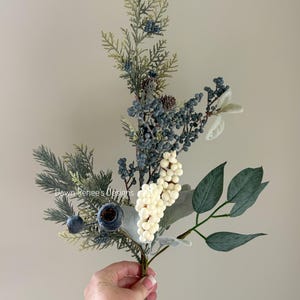 May include: A decorative floral stem featuring faux berries in white and blue, along with green leaves and pine needles. The stem is held in a hand and is against a neutral background. The stem includes a small pine cone.