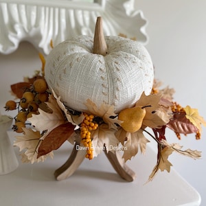 May include: A white fabric pumpkin with visible stitching detail sits atop a wooden stand, surrounded by artificial autumn foliage including tan maple leaves, small yellow berries, and a single golden pear, with the text Dawn Renee Designs.