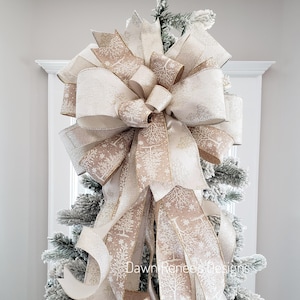 May include: A large, decorative Christmas tree bow made of cream and tan ribbons. The ribbons feature a winter tree pattern and gold trim. The bow is attached to a flocked Christmas tree, creating a festive holiday decoration.