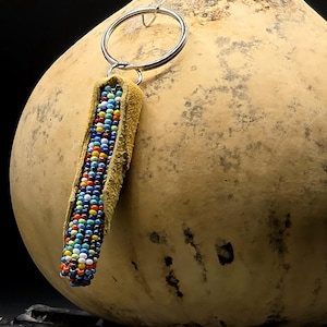 May include: A keychain featuring a cylindrical beaded ornament with a tan suede leather backing. The ornament is covered in small, multi-colored glass beads in shades of blue, red, yellow, and white, attached to a circular silver metal ring.