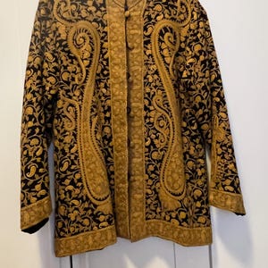 Può includere: Una giacca nera con intricati ricami floreali dorati. La giacca presenta un colletto alla coreana e un'abbottonatura frontale. Il ricamo dorato crea un motivo a forma di paisley sulla parte anteriore e sulle maniche. La giacca è appesa a un appendiabiti nero.