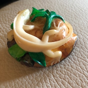 Pode incluir: Uma pequena figura de cerâmica de cor creme que representa um lagarto enrolado em torno de uma folha verde. A figura é detalhada com acentos marrons e amarelos.