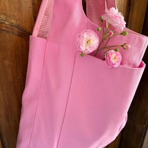May include: A light pink tote bag with a front pocket and a long strap. The bag is made of a sturdy fabric and is decorated with pink roses. The roses are placed in the front pocket of the bag.