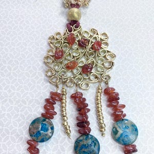 May include: A gold necklace with a large, intricate pendant featuring a swirling design and red carnelian stones. The pendant is attached to a burgundy cord and has multiple strands of carnelian and gold beads hanging from it.