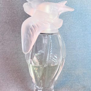 May include: A clear glass bottle of perfume with a frosted white bird-shaped stopper. The bottle has a faceted design and is filled with a clear liquid.