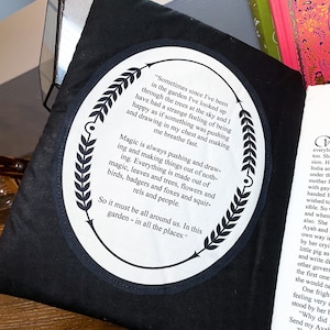 May include: A black fabric tote bag with a white circle patch featuring a quote about magic and nature. The quote reads, "Sometimes since I've been in the garden I've looked up through the trees at the sky and I have had a strange feeling of being happy as if something was pushing and drawing in my chest and making me breathe fast. Magic is always pushing and drawing and making things out of nothing. Everything is made out of magic, leaves and trees, flowers and birds, badgers and foxes and squirrels and people. So it must be all around us. In this garden - in all the places."