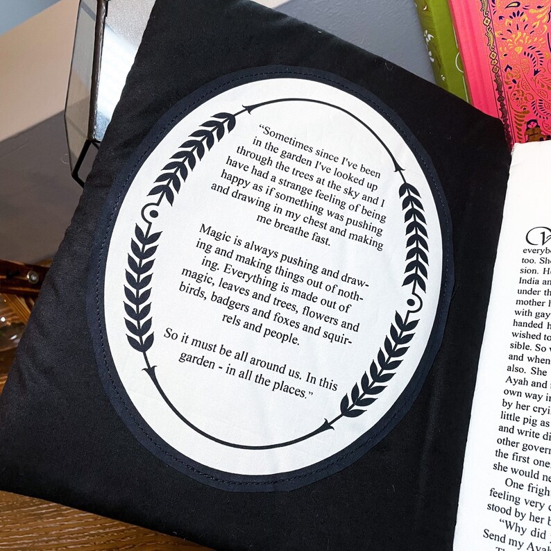 May include: A black fabric tote bag with a white circle patch featuring a quote about magic and nature. The quote reads, "Sometimes since I've been in the garden I've looked up through the trees at the sky and I have had a strange feeling of being happy as if something was pushing and drawing in my chest and making me breathe fast. Magic is always pushing and drawing and making things out of nothing. Everything is made out of magic, leaves and trees, flowers and birds, badgers and foxes and squirrels and people. So it must be all around us. In this garden - in all the places."