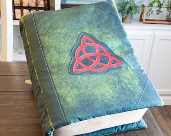 Puede incluir: Una almohada con forma de libro verde con un símbolo triquetra rojo en la parte delantera. La almohada está hecha para parecer un libro viejo y desgastado.
