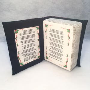 May include: A black and green book-shaped pillow with the text "Twas the night before Christmas..." and "'Twas the night before Christmas, when all through the house..." printed on the front and back covers. The pillow is made of fabric and has a soft, plush texture.
