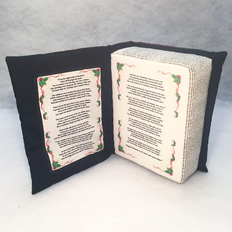 May include: A black and green book-shaped pillow with the text "Twas the night before Christmas..." and "'Twas the night before Christmas, when all through the house..." printed on the front and back covers. The pillow is made of fabric and has a soft, plush texture.