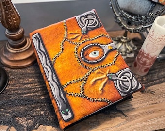 May include: A brown leather-bound journal with a stitched design. The cover features a large eye with a white iris and a black pupil, surrounded by snakes and stitching. The journal has a silver clasp and a silver snake design on the spine.