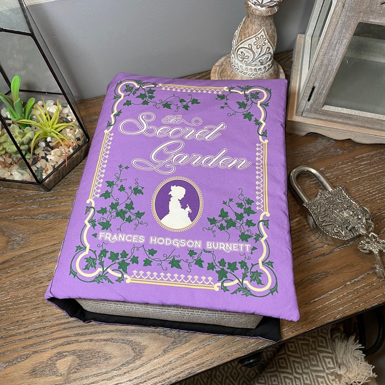 May include: A purple book cover with the title "The Secret Garden" and the author's name "Frances Hodgson Burnett". The cover features a decorative border of green ivy and a silhouette of a girl in a white dress.