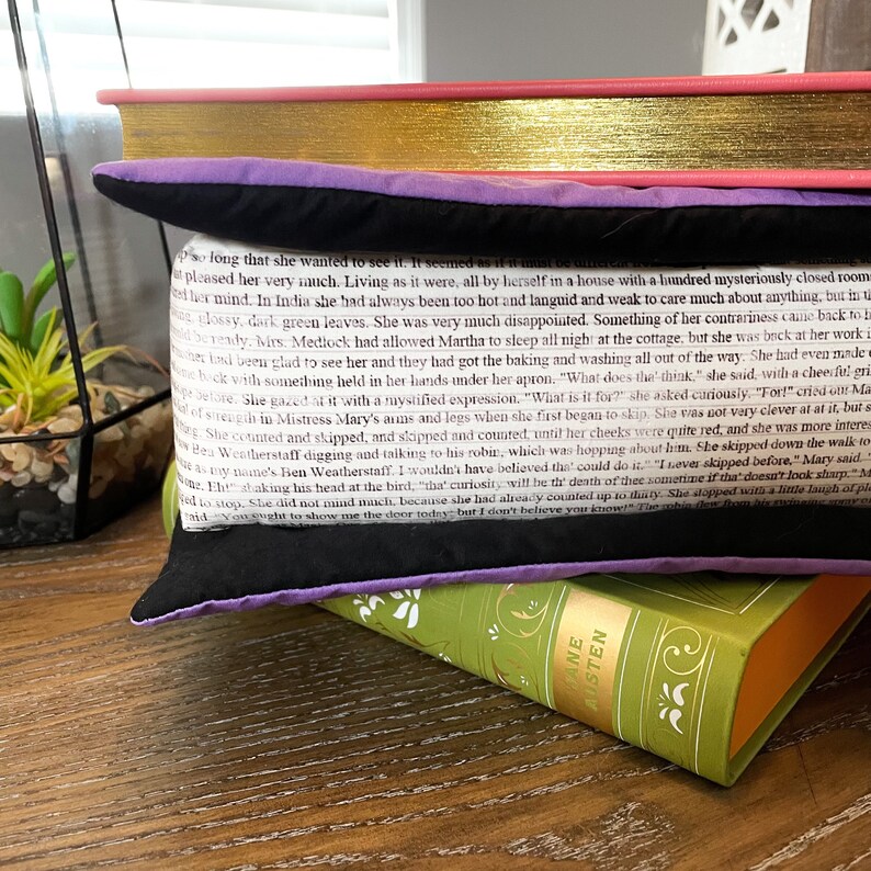 May include: A black and purple throw pillow with a white text overlay on a black background. The text is from a book and reads "so long that she wanted to see it. It seemed as if it innat ὡ&sigmaf; &alpha;&iota;&tau;&epsilon;ί&tau;&alpha;&iota; &sigma;&tau; pleased her very much. Living as it were, all by herself in a house with a hundred mysteriously closed room f her mind. In India she had always been too hot and languid and weak to care much about anything, but in glossy, dark green leaves. She was very much disappointed. Something of her contrariness came back to b berendy. Wirs. Mediock had allowed Martha to sleep all night at the cottage, but she was back at her work uner band been glad to see her and they had got the baking and washing all out of the way. She had even unde we bock with something held in her hands under her apron, "What does the think she said with a cheerful cri Not Delote. She gazed it with a mystified expression, "What is it for?" she asked cariously, "For!" cried our Ma of strength in Mistress Mary's auns and legs when she first began to skap, She was not very clever at at it, but s She counted and skipped, and slipped and counted until her cheeks were quite red, and she was more interes Cow Ben Weatherstaff digging and talking to his robin which was hopping about him. She skipped down the walleto asy-Baane's Ben Weatherstaff. I wouldn't have believed the could do it. I ever skipped before." Mary said te. Eht with a little laugh of aid You wught to show me the door today, but I don't believe you know! The robin hơn b&aacute;c ruinning shaking his head at the bird. that curiosit will be the death of thee sometime if that doesn't look sharp M"