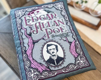 Op de afbeelding: Een grijs en roze decoratief kussen met een vintage boekomslagontwerp met een portret van Edgar Allan Poe en de tekst "Edgar Allan Poe His Complete Tales & Poems".