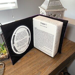 May include: A purple and black book pillow with a white fabric cover and a quote about magic on the front. The book is open to reveal a page with the text "Chapter 1 There is No One Left".