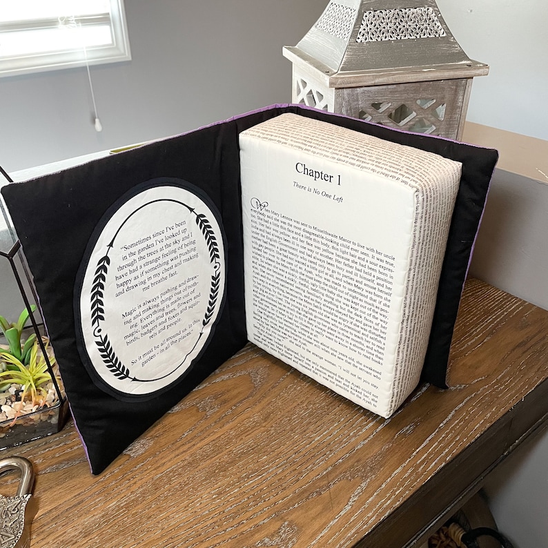 May include: A purple and black book pillow with a white fabric cover and a quote about magic on the front. The book is open to reveal a page with the text "Chapter 1 There is No One Left".