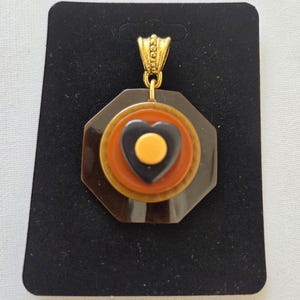 May include: A vintage pendant with a gold-colored bail. The pendant features a dark brown, octagonal base, with concentric circles of gold, orange, and a black heart shape with a yellow center. The pendant is displayed on a black card.