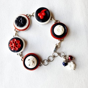 May include: A silver-toned charm bracelet featuring a series of decorative buttons in red, white, and black. The buttons have various floral and abstract designs. The bracelet is accented with dangling charms.