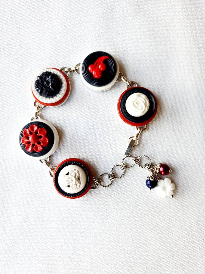 May include: A silver-toned charm bracelet featuring a series of decorative buttons in red, white, and black. The buttons have various floral and abstract designs. The bracelet is accented with dangling charms.