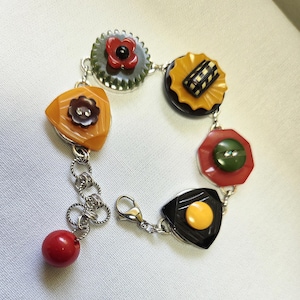 May include: A vintage-style bracelet featuring a series of colorful, decorative buttons in various shapes and sizes. The bracelet includes orange, red, yellow, black, and green buttons, connected by a silver chain. A red bead dangles from the end.