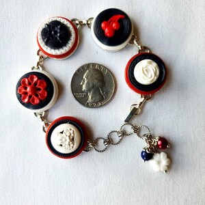 May include: A vintage-style bracelet featuring a series of decorative buttons in red, black, and white. The buttons have various embossed designs, including flowers and cherries. The bracelet is connected by silver-toned chain links and has small charms.
