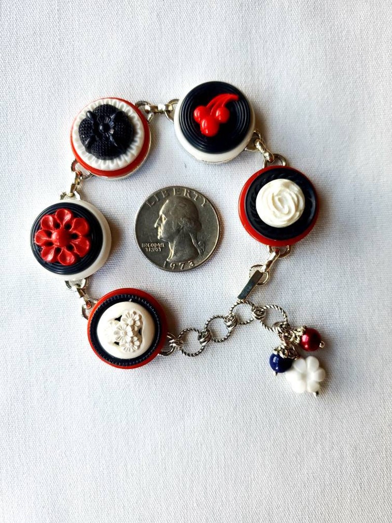 May include: A vintage-style bracelet featuring a series of decorative buttons in red, black, and white. The buttons have various embossed designs, including flowers and cherries. The bracelet is connected by silver-toned chain links and has small charms.