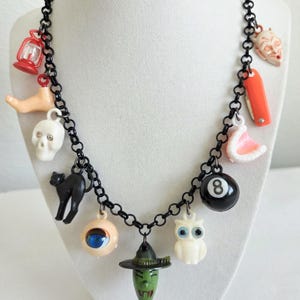 May include: A black chain necklace with various Halloween-themed charms. Charms include a skull, black cat, eyeball, witch, owl, eight ball, devil face, and a red lantern. The necklace is displayed on a white mannequin.