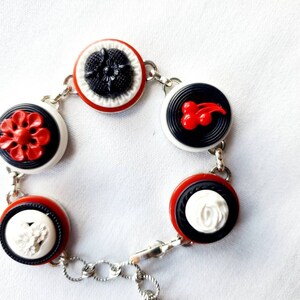May include: A bracelet featuring a series of decorative buttons in red, black, and white. The buttons are connected by a silver chain. The buttons have various designs, including flowers and cherries.