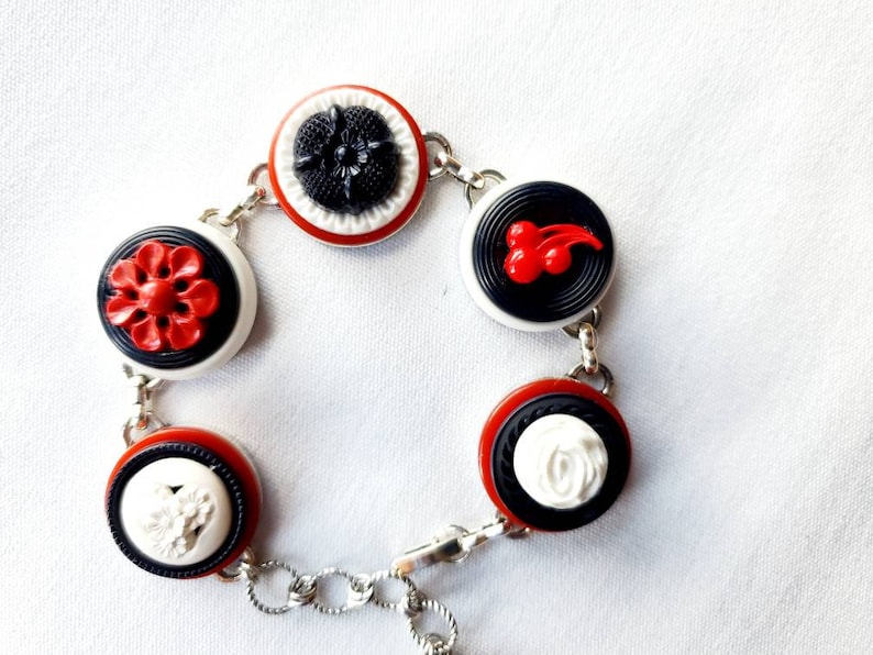 May include: A bracelet featuring a series of decorative buttons in red, black, and white. The buttons are connected by a silver chain. The buttons have various designs, including flowers and cherries.