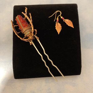 Puede incluir: Una horquilla dorada con un dise&ntilde;o de escarabajo rojo y verde. El escarabajo tiene un dise&ntilde;o detallado con acentos rojos y verdes. La horquilla est&aacute; sobre un fondo negro. La imagen tambi&eacute;n muestra un par de pendientes dorados con colgantes en forma de hoja.