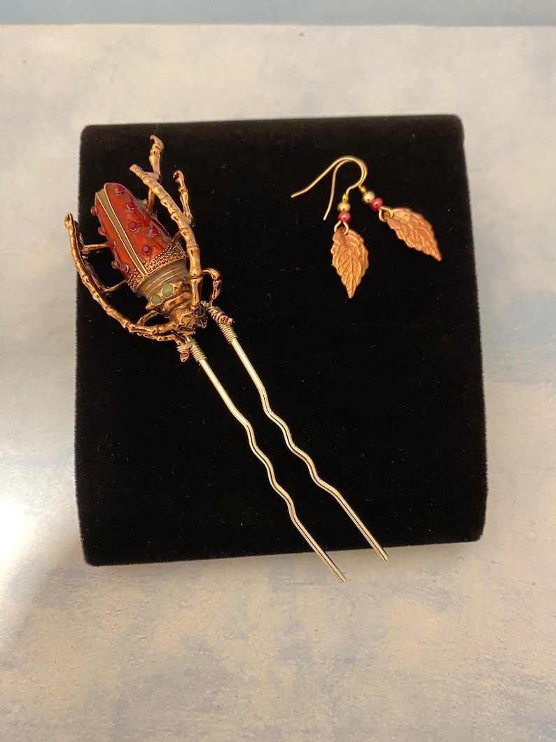 Puede incluir: Una horquilla dorada con un dise&ntilde;o de escarabajo rojo y verde. El escarabajo tiene un dise&ntilde;o detallado con acentos rojos y verdes. La horquilla est&aacute; sobre un fondo negro. La imagen tambi&eacute;n muestra un par de pendientes dorados con colgantes en forma de hoja.