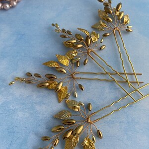 May include: Three gold hair pins with delicate gold leaf and bead embellishments. The pins are arranged in a fan shape on a light blue background.