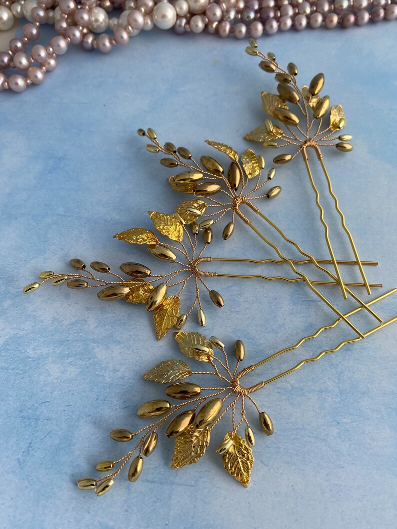 May include: Three gold hair pins with delicate gold leaf and bead embellishments. The pins are arranged in a fan shape on a light blue background.