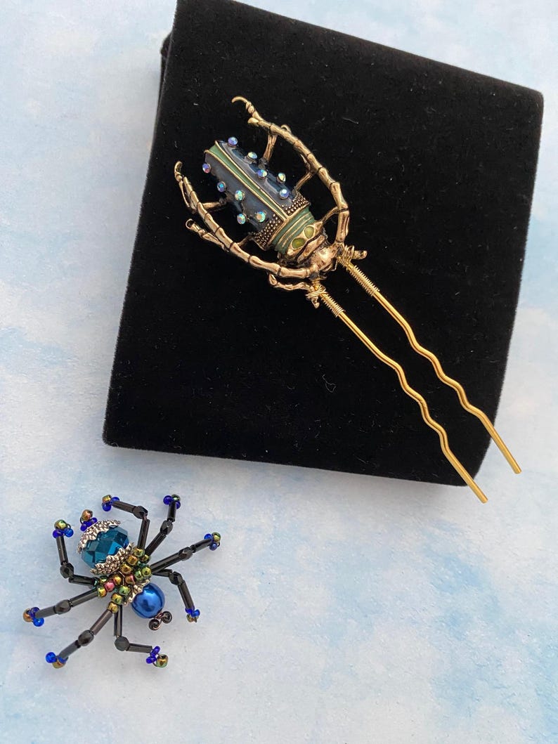 May include: Two hair clips featuring insect designs. One clip is a gold-toned beetle with blue and green enamel and rhinestones. The other clip is a black beaded spider with a blue gemstone in the centre.